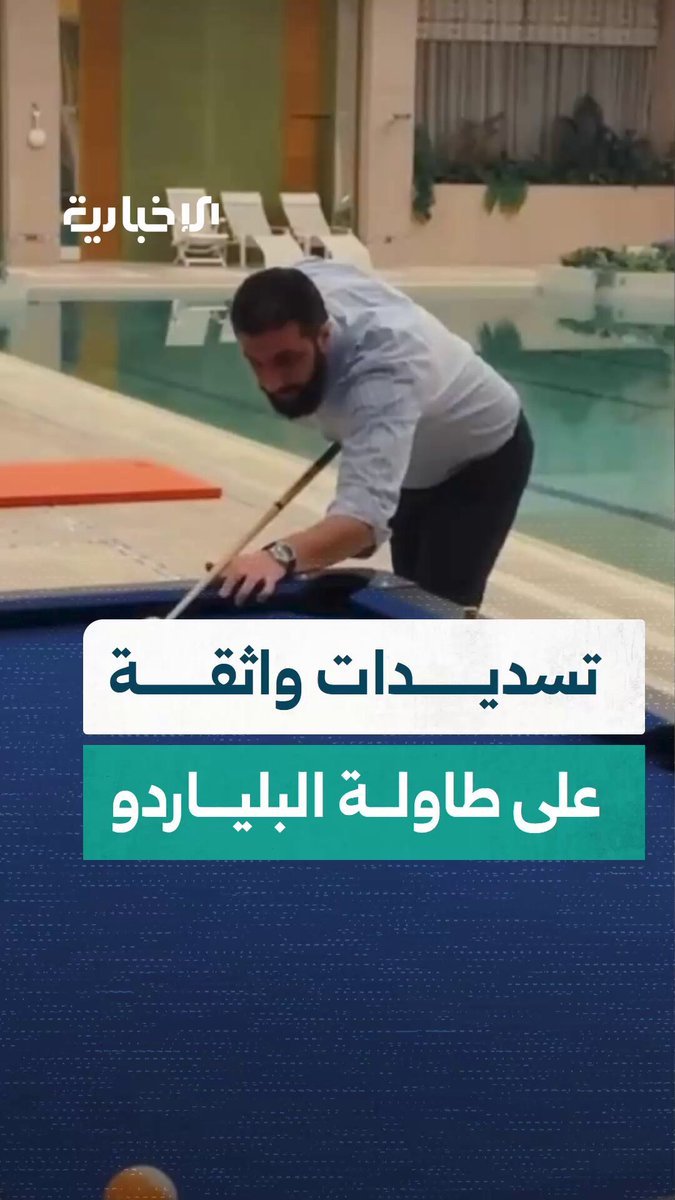 A man in a light blue shirt and dark pants plays billiards on a blue pool table near an indoor swimming pool with clear water. The setting features a modern room with wooden paneling, lounge chairs, and large windows. Arabic text overlays the video, including "الرئيس الشرع" and other phrases. The man uses a cue stick to strike balls on the table, focusing intently on his shots.